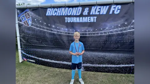 A boy with blond hair, wearing a football blue jersey and black shorts, long socks and white trainers. He is standing in front of a large black banner. He is wearing a medal around his neck.