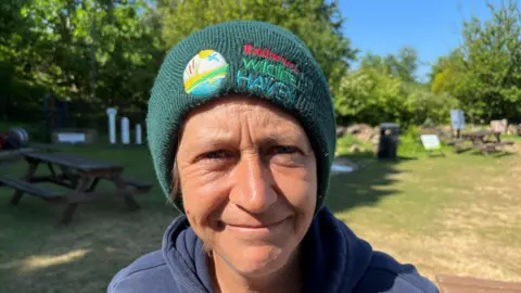 John Devine/BBC Becky White, 45, has a green bobble hat on and blue fleece. In the background are picnic benches and greenery.
