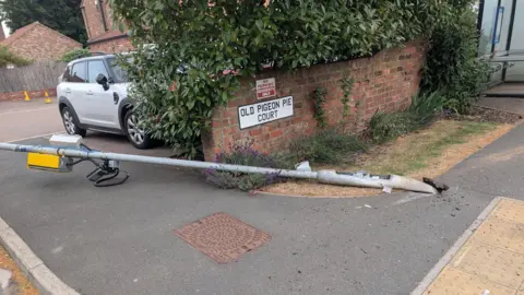 A yellow box attached to a lamp-post is pictured bent over and lying almost horizontally on the ground.