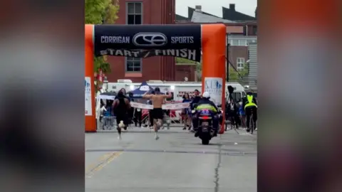 A runner with his hands raised and another runner coming up behind him 