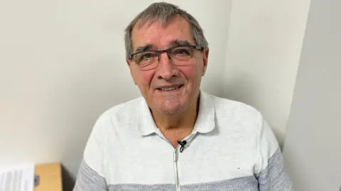 Alex looks directly at the camera, smiling, he has short grey hair with flecks of white and wears black wire framed glasses with a clear base to the frame. He is wearing a light grey and darker grey knitted polo top with a golden zip up to the collar.