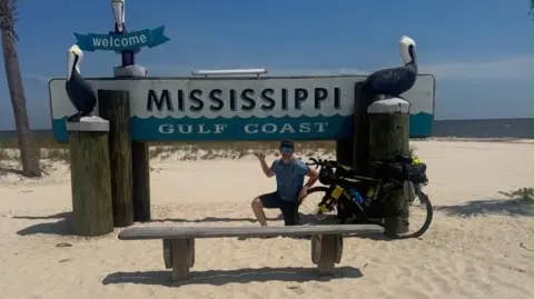 Joe Dick A man in a light blue England football shirt and black shorts kneels in front of a sign on a beach which says Mississippi Gulf Coast. The sign has two wooden Pelicans and says welcome. The man's bike is propped up against the sign.
