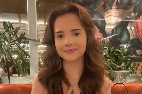 Freya Price Freya is sat in a restaurant, she has long brown hair and is smiling at the camera. She is wearing an amber pendant. She is sat on an orange leather sofa and is there are plants behind it. 