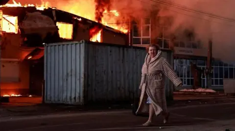 A woman walks past a burning house at the site of Russian missile and drone strike, amid Russia's attack on Ukraine, in the town of Brovary, in Kyiv region