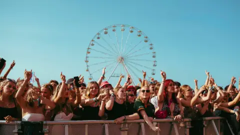 sammcmahon_@kiwimcmc A crowd of festivalgoers stands at the front barrier with arms raised while a large Ferris wheel rises in the background against a clear blue sky.