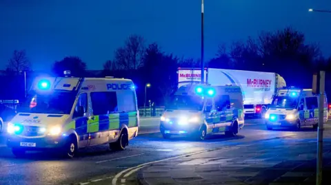 A convoy of police vans driving along a road. Their blue lights are illuminated in the dawn gloom.