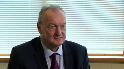 BBC A man with grey hair in a navy suit, white shirt and burgundy tie