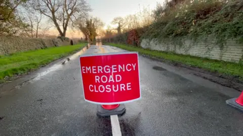 BBC Emergency Road Closure sign on a road with grass verges either side.