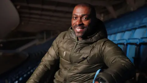 Prostate Cancer UK A man with dark hair is sitting in stadium with rows of blue plastic seats behind him. It's at night, and he's wearing a khaki green puffer jacket