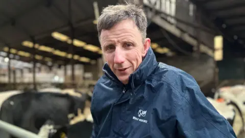 An image of Mervyn Gordon. He is a middle-aged man with greying hair. He is standing in front of a herd of cows but the background is blurred. He is wearing a navy raincoat and is looking at the camera with a neutral expression on his face.
