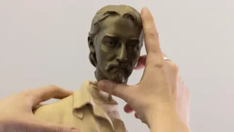 A pair of hands attach a head to the neck of a small white statue. The head is a man with a moustache.