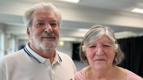 Trevor has short white hair and a grey beard, and is wearing a white polo top. Lorraine has shoulder length grey hair and is wearing a pink top. 