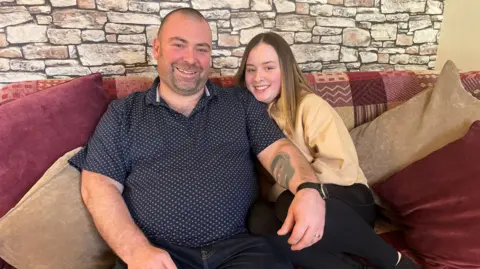 Steve Braid and his daughter Niamdh sit close together on a pink sofa, smiling at the camera. Steve is wearing a navy short-sleeved shirt with white dots and dark jeans. Niamdh is wearing her beige "proud to be deaf" hoodie and is snuggled close to her dad. A wall of stone-effect wallpaper is behind them.