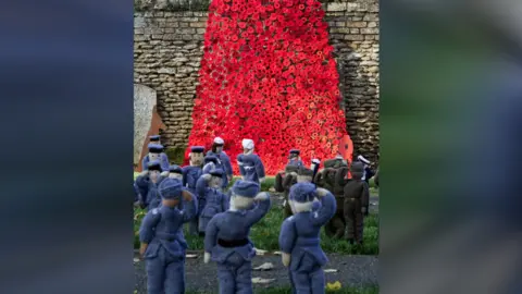 Mandy Wright Photography A waterfall of red poppies on a stone wall with small knitted soldiers on the grass in front of it 