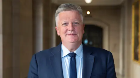 A man with short grey hair in a navy suit with a blue shirt and tie