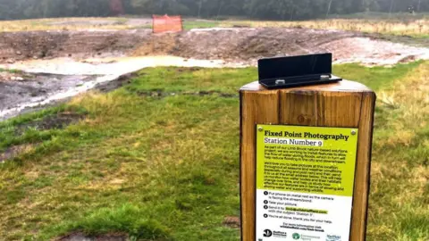Sheffield and Rotherham Wildlife Trust An outdoor scene shows an expanse of grass with a large ditch towards the top of the picture. A small orange barrier fences of one small section of the edge of the ditch. In the foreground stands a wooden post with a notice attached. The background of the notice is green from the top to halfway down and then the background is white. There is black lettering in on the notice. At the top it says Fixed Point Photography in large letters with Station Number 9 written underneath. The rest of the lettering is too small to read.