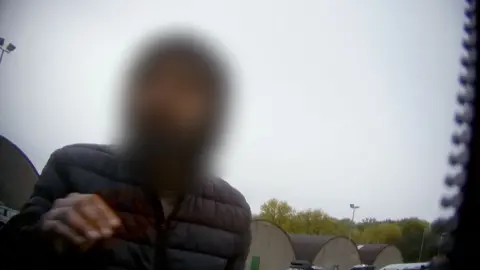 Man with a blurred face wearing a black anorak. There is grey sky behind, with trees in the distance. The image was taken undercover, and part of a zip is visible on the right hand side.