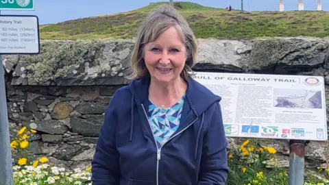 Maissie Presslie A woman with medium length, blondish brown hair wearing a blue zip-up top with a patterned blouse underneath. She is standing in front of a sign for the Mull of Galloway trail with a wall and field and flowers behind her.
