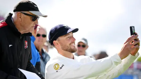 Getty Images Ollie Robinson in cricket whites and a navy cap smiling as he takes a selfie with a man in black hoody, cap and sunglasses behind him. 