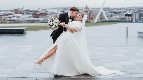 Mark Hamill Photography Caoimhe and Ollie are in front of the peace bridge in Derry. He is dipping her and her leg is up showing her white wedding shoes. She is wearing a white satin wedding dress that is off the shoulder. She is holding a flower bouquet and a glass of champagne. 