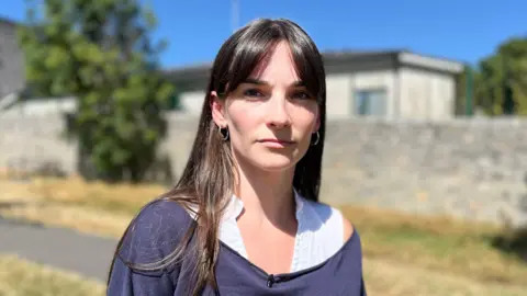 A young woman wearing a dark purple jumper over a white blouse stands outside on a sunny day, She has long brown hair and behind her a stone building is visible, but not in focus