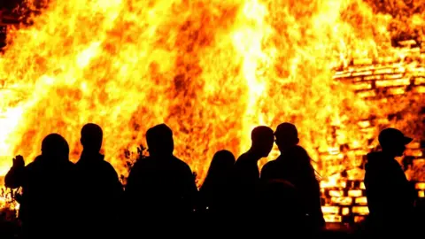 PA Media A group of silhouettes, look like young people, standing in front of a lit bonfire, with lots of emblems across the pallets and billows of smoke.