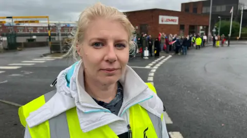 The South West Regional Officer for Unite, Shevaun Hunt wearing a high vis vest and white coat facing the camera. She has blonde hair tied back and is standing outside the front gates of the Yeovil factory. 