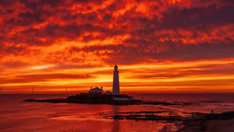 A lighthouse on a rocky outpost off the coast of Whitley Bay, surrounded by the sea. It stands dark against a dramatic, orange sunset in a sky which is flecked with red-streaked clouds.