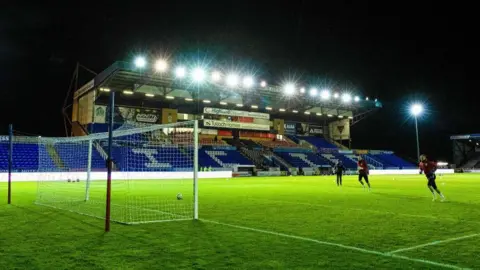 SNS The lights in the main stand are on, illuminating the pitch where some players are training in front of one of the goals.