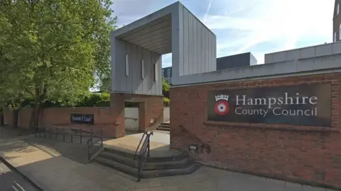 A brick wall with a sign saying 'Hampshire County Council' with its crown and rose logo. There are three steps up to an opening in the wall.