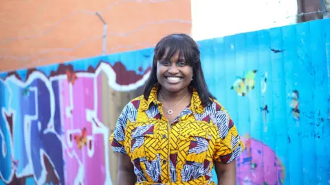 Louise Ndibwirende Ms Ndibwirende in a brightly-coloured yellow dress standing, smiling, in front of a fence which has street art painted on it.