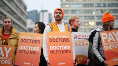 PA Media Resident doctors wearing orange beanie hats and carrying orange placards bearing the wears Doctor Need Jobs