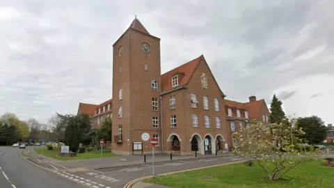 Google Exterior of Spelthorne Borough Council offices in Staines. A small tree in the foreground. There is are no entry signs to a road just outside the building