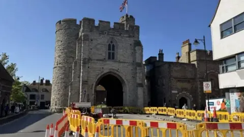 A stone tower with a large archway underneath it, flying a union jack flag from its turrets. Road access to the archway is blocked by orange traffic cones and yellow plastic barriers.