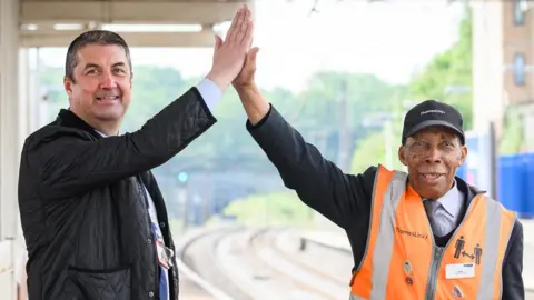 Joe Healy (left) and Siggy Cragwell (right) sharing a high five. Joe is wearing a black coat over a shirt and tie. Siggy is wearing a high-vis orange vest. They are on a station platform, with greenery visible in the background.