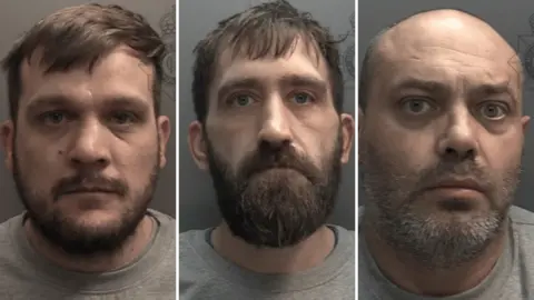 Three men wearing grey sweatshirts are staring into a camera with a grey wall behind them. All three have beards, with the first two also having short, dark hair and the third a bald head.