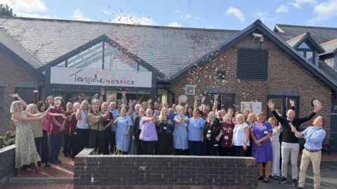 A crowd of staff stand outside the single-storey Teesside Hospice building and let off confetti streamers while cheering. Lots of staff have their hands held in the air and they are all smiling. Jets of confetti are floating in the air.