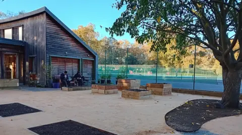 A general view of the new sanctuary garden in the process of being completed. There are two people sitting on a bench. There's a tree in the right hand corner and a sports court in the background.