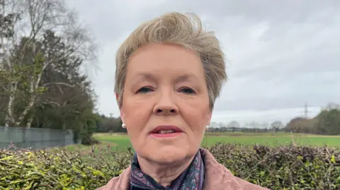 A woman with short fair hair standing in front of a hedge with fields in the background. She is wearing a light brown top and paisley scarf.