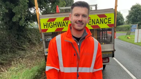 BBC/ Ian Reeve Keane Duncan, from North Yorkshire Council, wears an orange hi-vis jacket and stands in front of a highway maintenance vehicle