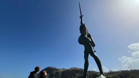 The picture shows a huge seaside statue of a woman holding a sword straight up into the air. The figure stands on a plinth beside the water, with dramatic cliffs rising behind her. The sky is bright and clear, with a few clouds drifting over.
Two people are standing in the bottom left corner of the frame, looking up at the sculpture. 