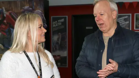 Derian House handout Caroline Taylor, of Derian House, with long blond hair wearing a white jumper is smiling as she chats to Steve Pemberton who is wearing a navy coat and beige t-shirt. 