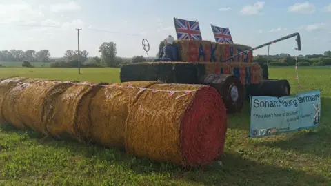 Soham Young Farmers A straw bale sculpture showing a combine harvester. On the right are round bales and behind it is the combine shape, with a figure made from a stuffed pair of blue overalls. On its side it says Back British Farming in blue and red letters. On its roof are two Union flags. It is resting in a field and in front is a sign saying Soham Young Famers.