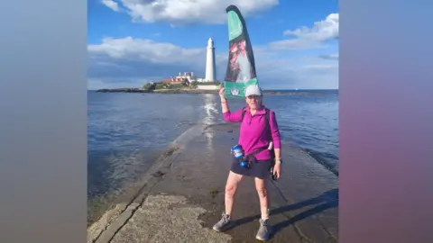 Susan Dray A woman in sportswear standing on the causeway to St Mary's Island, flooded due to the tide, with the white lighthouse and cottages of the island vicible behind her under a bright and cloudy sky. She is wearing sportswear - a bright pink fleece, dark shorts, trainers and a blue baseball cap with a collection tin hanging from her waist. A flag or banner with a picture of a young man can be seen behind her.