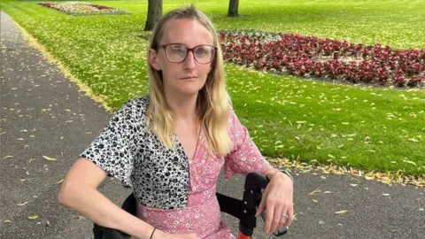 Laura Earl is sitting in a wheelchair and looking at the camera with a neutral expression on her face. She is in a park, on the grey tarmac path, with a grassy area and red flower bed behind her. She has long blonde hair, glasses and is wearing a pink flowery dress.