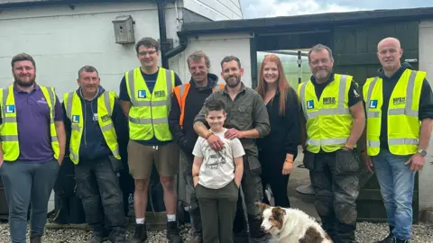 A young boy stands with his parents and a dog in front of a house. Six men in hi-vis jackets stand alongside them. They are all smiling at the camera.