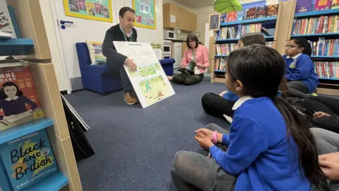 Children sat in a school library. They are crossed legged and looking at a man reading a story board.