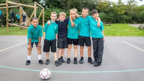 Cabot Learning Federation Six primary school pupils stand together on a playground with their arms around each other. Most are wearing turquoise polo shirts and dark shorts or trousers, with a football resting on the ground in front of them and a climbing frame behind.