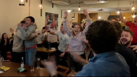 Pub-goers are celebrating among tables and chairs in a pub. Some are hugging and some are waving their hands in the air.