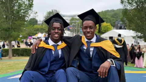 Two young men in Navy blue suits wearing black and yellow robes and black mortarboards 